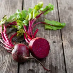 Zwei frische rote Bete, eine davon angeschnitten, liegen mit grünen Blättern auf Holz. Perfekt, um rote Beete roh zu essen.